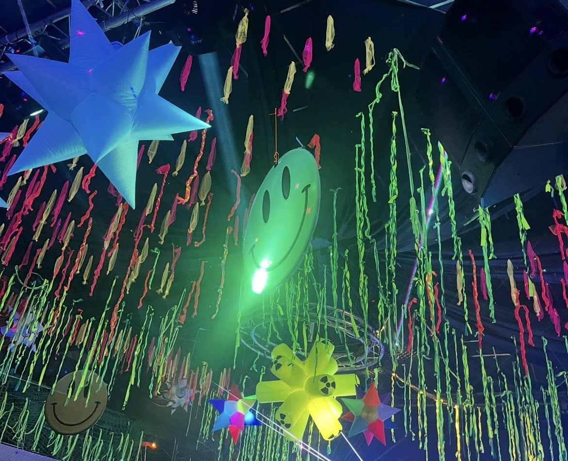 Bright neon decorations hanging from a ceiling, including a glowing smiley face and colourful streamers, representing sensory overload and overstimulation