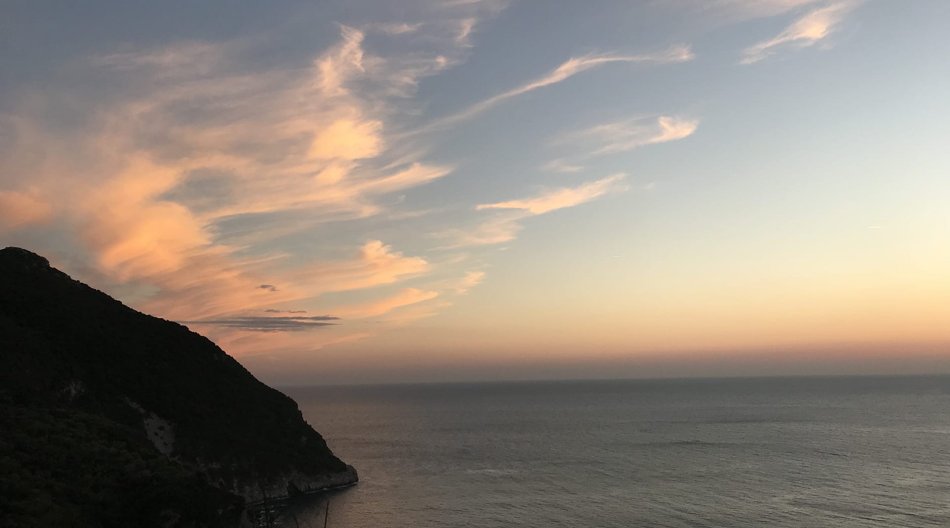 Soft pastel clouds over a calm sea at sunset beside a dark headland.